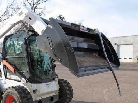 A Bobcat skid steer, with its wide bucket lowered and a powerful hammer attachment at the ready, sits idle in a parking lot.