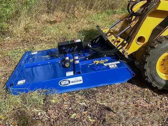 The image shows a yellow skid steer with a blue mower attachment in a field. The tractor is parked on a dirt path, and the mower is raised off the ground. 