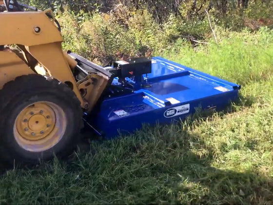  The image shows a mower attached to a yellow skid steer driving through a field of tall grass. The grass is green and appears to be freshly cut.