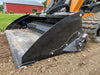 A close-up of a skid steer bucket with a worn-out cutting edge, resting on a pile of dirt in front of a red barn.