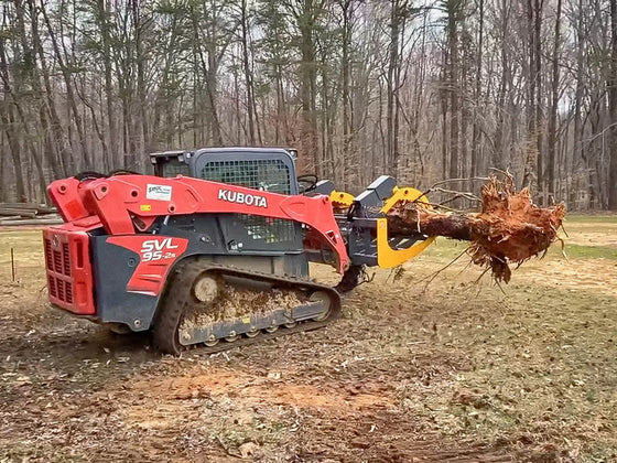 A Kubota skid steer loader with a grapple rake attachment is parked on a pile of wood chips. The grapple rake is attached to a tree stump that has been ground into mulch.
