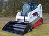 A skid steer loader with a hydra bucket attachment, parked on a lush green field.