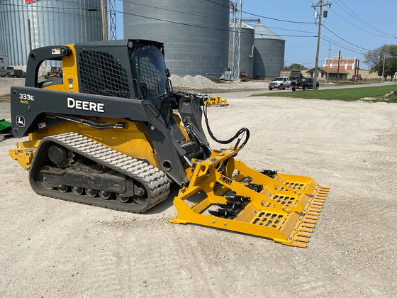 A John Deere skid steer loader with a land leveler attachment parked on a dirt road.