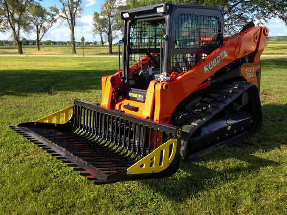  A Kubota SVL90 skid steer loader with an Rock bucket attachment parked on a grassy field. The skid steer loader has its front bucket raised and is turned slightly to the right. The grass is green and there are a few trees in the background.