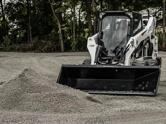 The image shows a skid steer with a large bucket on top of a pile of gravel. The skid steer is white and black, and the bucket is black. The pile of gravel is in a vacant lot, and there is a row of trees in the background.