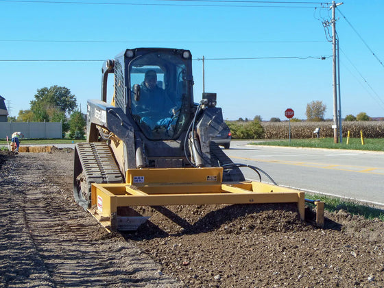 A skid steer with a DoMor SSF-72 box grader attachment, parked on a gravel surface. The attachment is yellow with black branding and has an open-box design.