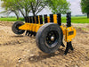 A yellow tracked skid steer loader with black rubber tracks and a yellow plow attachment parked on a dirt construction site. The plow attachment has six curved teeth and tapers to a point at the front. Behind the skid steer loader is a pile of dirt and a blue dumpster. 