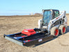 A skid steer loader equipped with a brush mower attachment working on a barren field, highlighting the mower's broad cutting deck and the skid steer's muddy wheels, against a backdrop of a clear sky and distant farm structures.