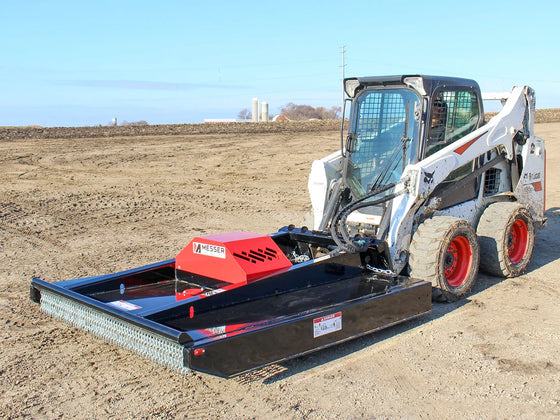 A skid steer loader equipped with a brush mower attachment working on a barren field, highlighting the mower's broad cutting deck and the skid steer's muddy wheels, against a backdrop of a clear sky and distant farm structures.