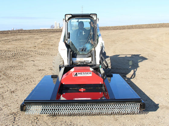 Frontal view of a skid steer equipped with a red and black brush mower attachment, positioned on a plowed field, with the operator visible in the cabin, ready to begin land clearing work.